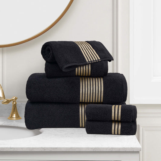 A stack of Melissa Bath Towel Set, made from 100% cotton towels with gold stripes, is neatly arranged on a white marble bathroom countertop beside a gold faucet and mirror.
