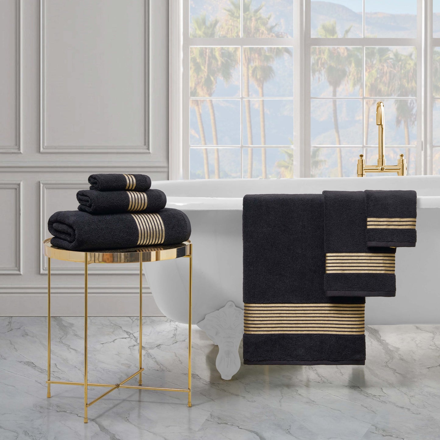 A white bathtub with a gold faucet and marble floor is accented by dark gray Melissa Bath Towel Set towels—100% cotton, zero-twist weave, gold-striped—folded on a gold side table and draped over the tub.