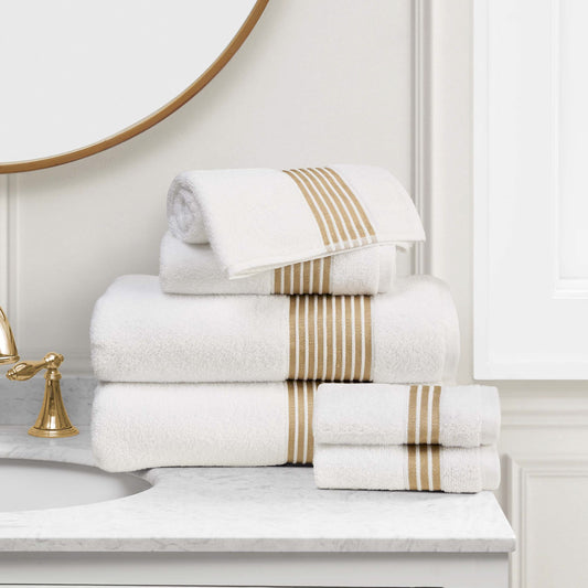 A stack of Melissa Bath Towel Set with beige stripes is neatly arranged on a marble bathroom countertop beside a gold faucet and a round mirror.