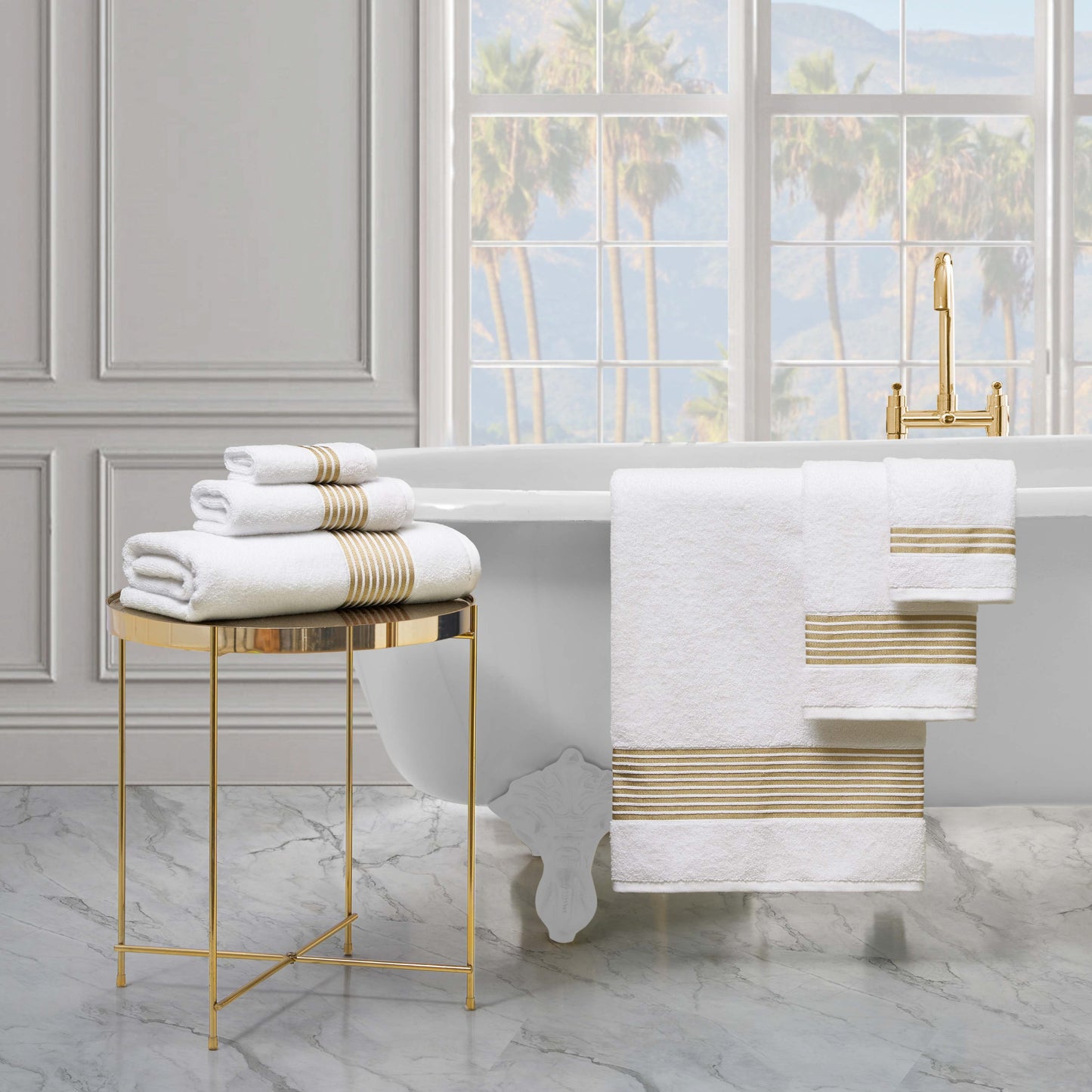 A luxurious bathroom with a white clawfoot tub, gold fixtures, and a marble floor features the Melissa Bath Towel Set—plush 100% cotton towels with gold stripes—neatly folded on a gold table and draped over the tub. Palm trees are visible through large windows.
