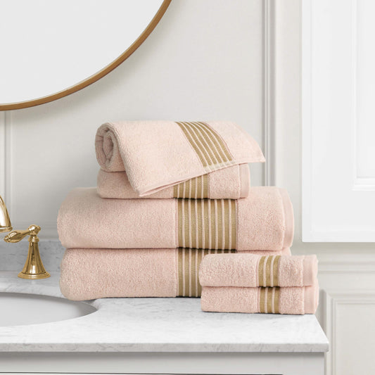 A neatly folded Melissa Bath Towel Set in light pink with beige stripes sits on a marble bathroom countertop next to a gold faucet and round mirror.