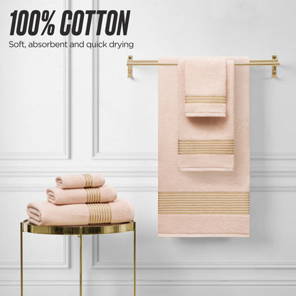 The Melissa Bath Towel Set in soft pink with beige stripes is displayed on a gold rack and table against a white wall. Text reads: 100% Cotton – Soft, absorbent, and quick-drying zero-twist towels.
