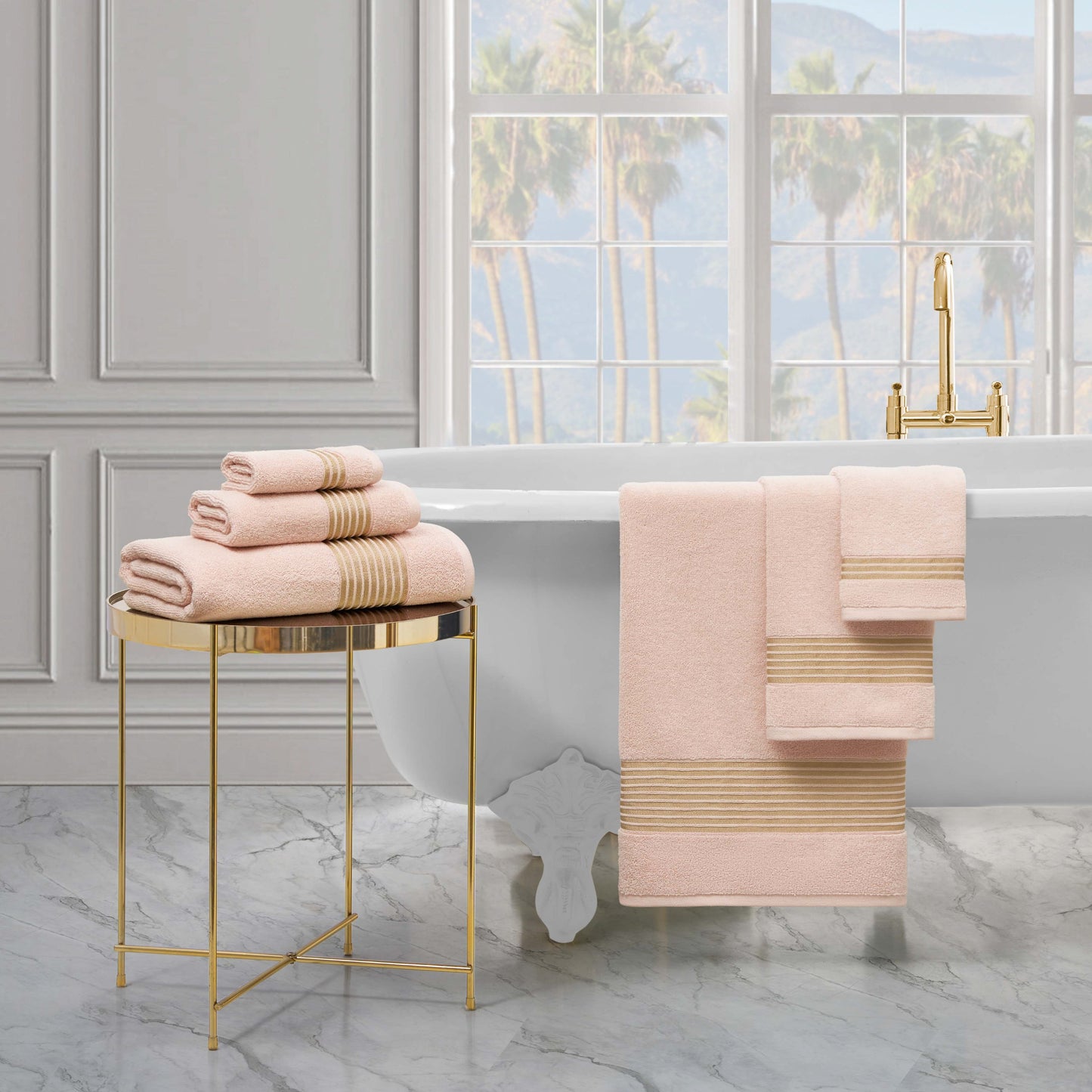 A luxurious bathroom with a freestanding white tub, gold fixtures, and a stacked Melissa Bath Towel Set on a gold side table, plus large windows revealing palm trees and mountains in the background.