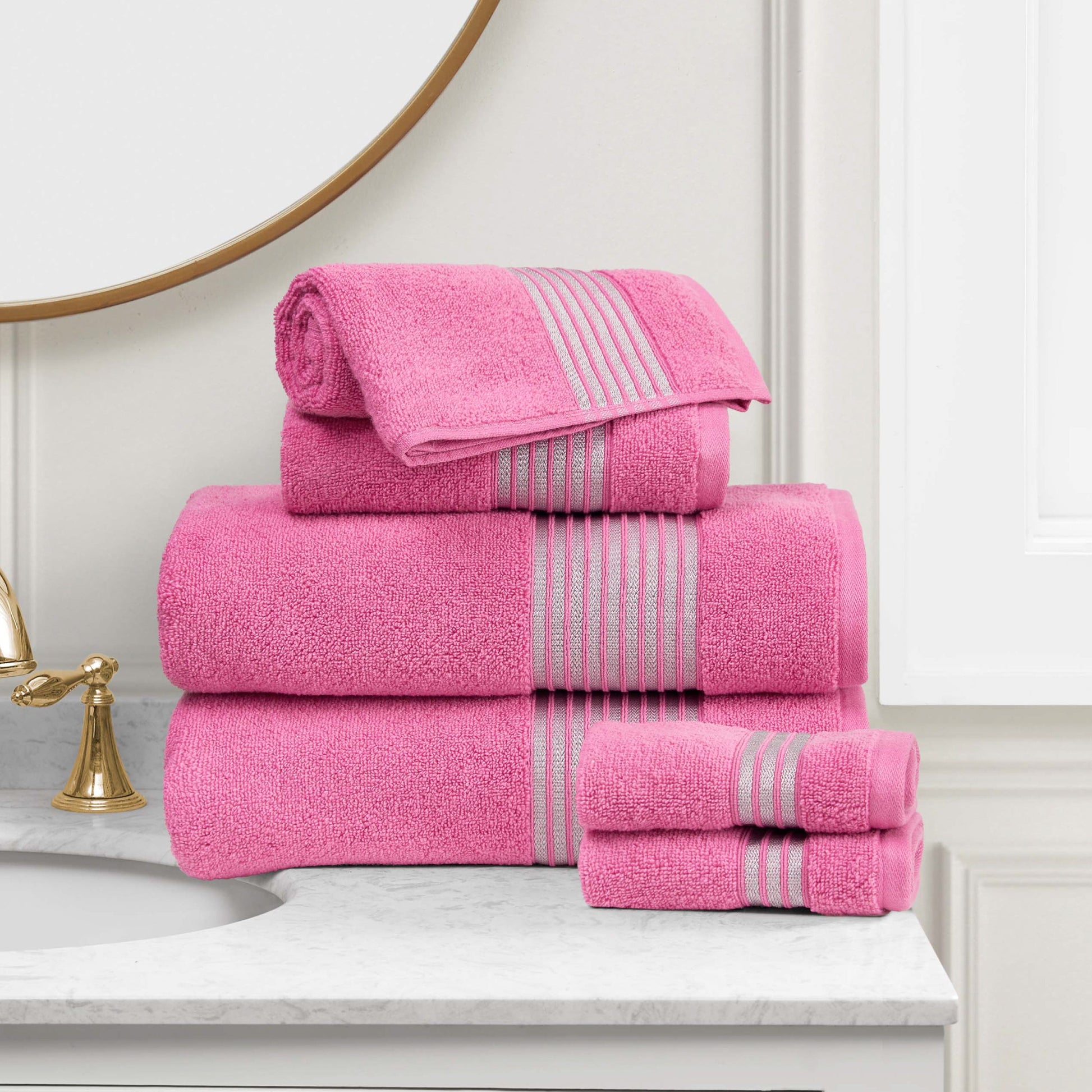 A stack of pink Melissa Bath Towel Set with gray stripes is neatly arranged on a marble bathroom counter beside a gold faucet and round mirror.