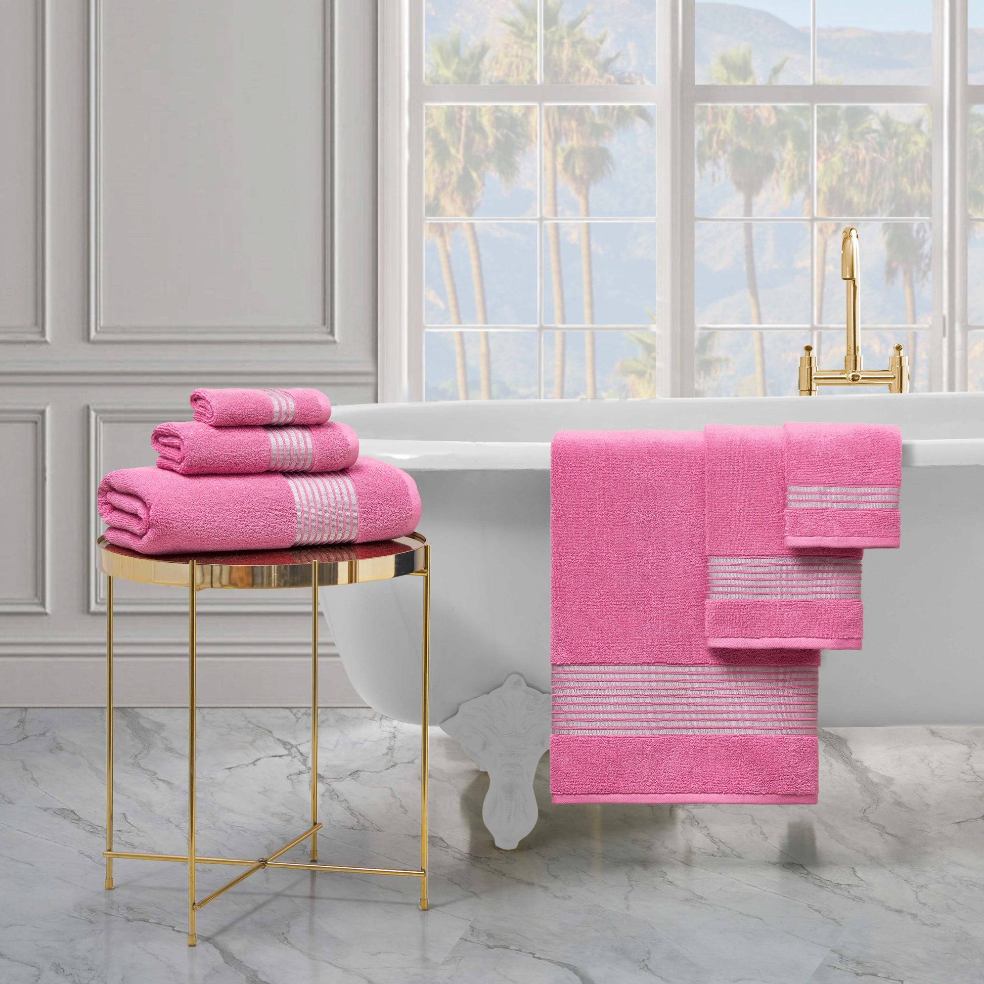 A luxurious bathroom features a white clawfoot tub, a gold side table with the Melissa Bath Towel Set, and matching pink towels hanging on the tub. Large windows offer views of palm trees outside.