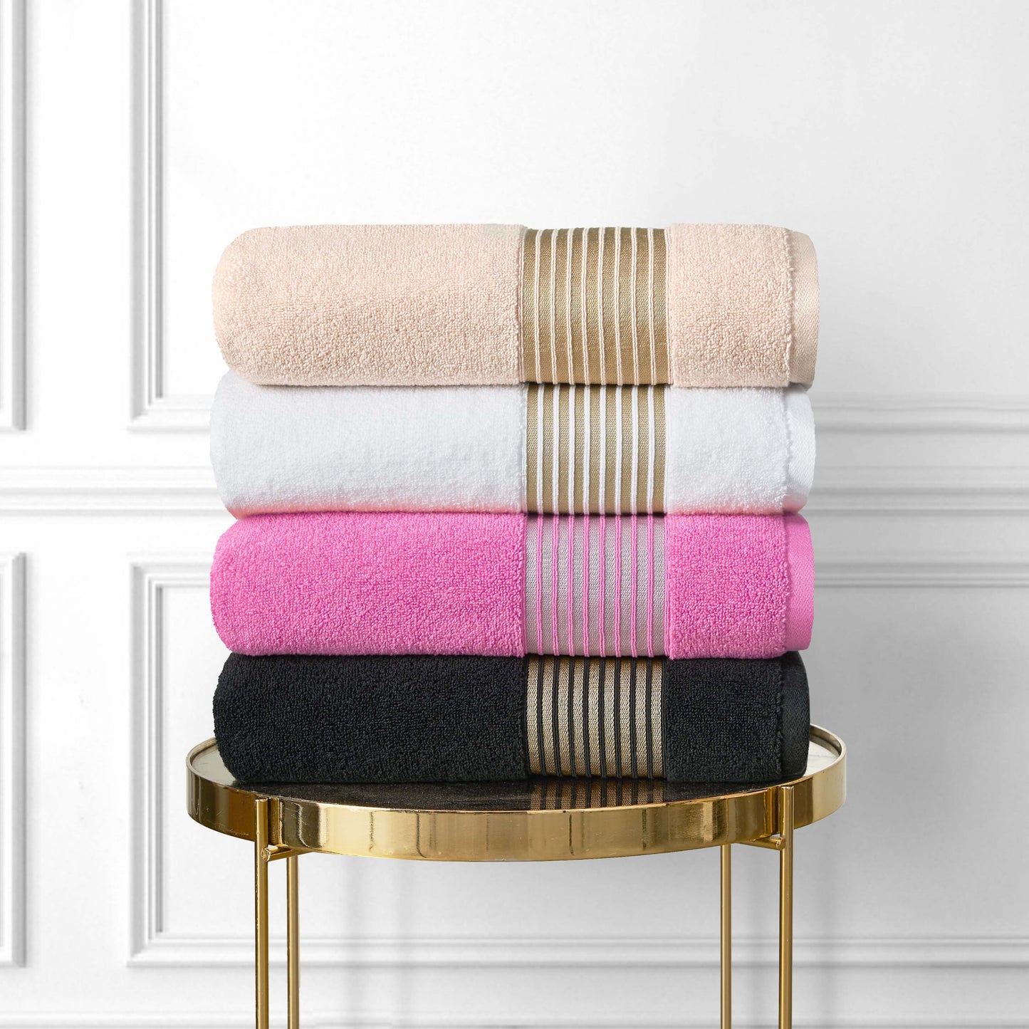 The Melissa Bath Towel Set, with four neatly folded 100% cotton towels in beige, white, pink, and black—each stacked on a gold round table—features beige and black zero-twist towels accented with elegant gold stripes.