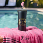 A Couture Hydration Stainless Steel Tumbler Cup from the 6-Piece Set, featuring a gold logo and pink bear topper, rests on a pink Juicy towel by the pool with palm trees and sparkling water in the background.