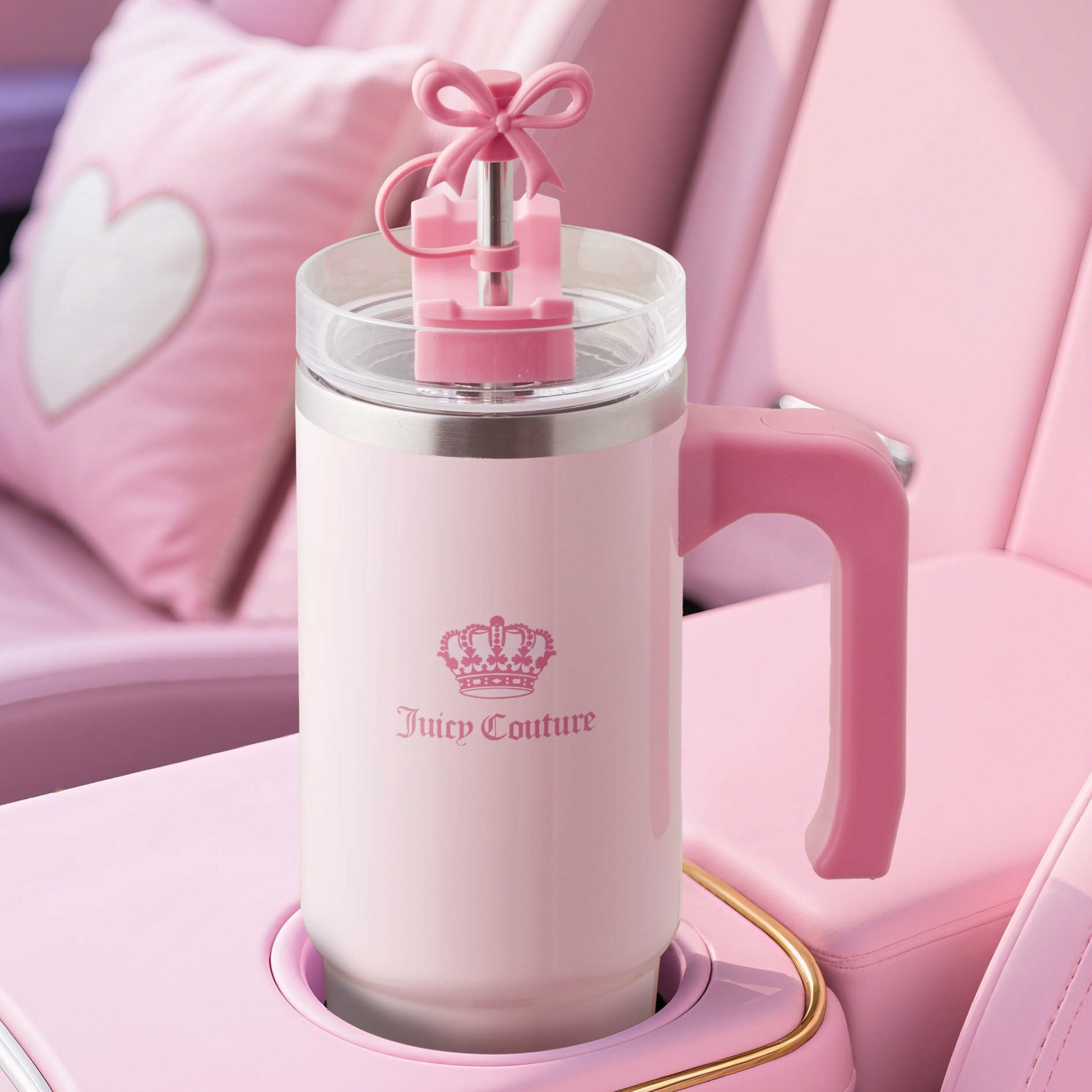 A large pink and white Jetsetter Hydration Stainless Steel Tumbler with a handle sits in a pink car cup holder. The straw has a pink bow, and a heart-shaped pillow can be seen on the seat in the background.