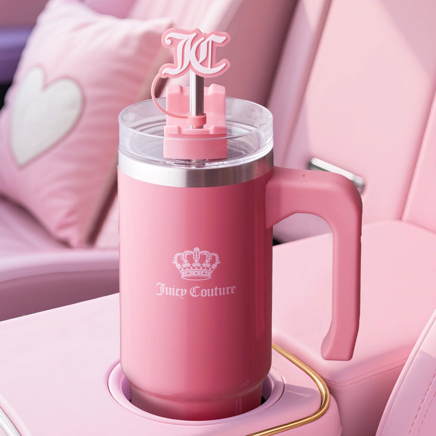 A pink Jetsetter Hydration Stainless Steel Tumbler with handle, clear lid, “JC” logo straw, and neoprene crossbody pouch sits in a pink car’s cup holder with a heart pillow in the background.