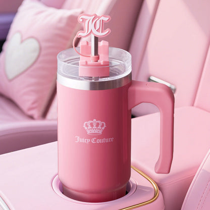 A pink Jetsetter Hydration Stainless Steel Tumbler with handle, clear lid, “JC” logo straw, and neoprene crossbody pouch sits in a pink car’s cup holder with a heart pillow in the background.
