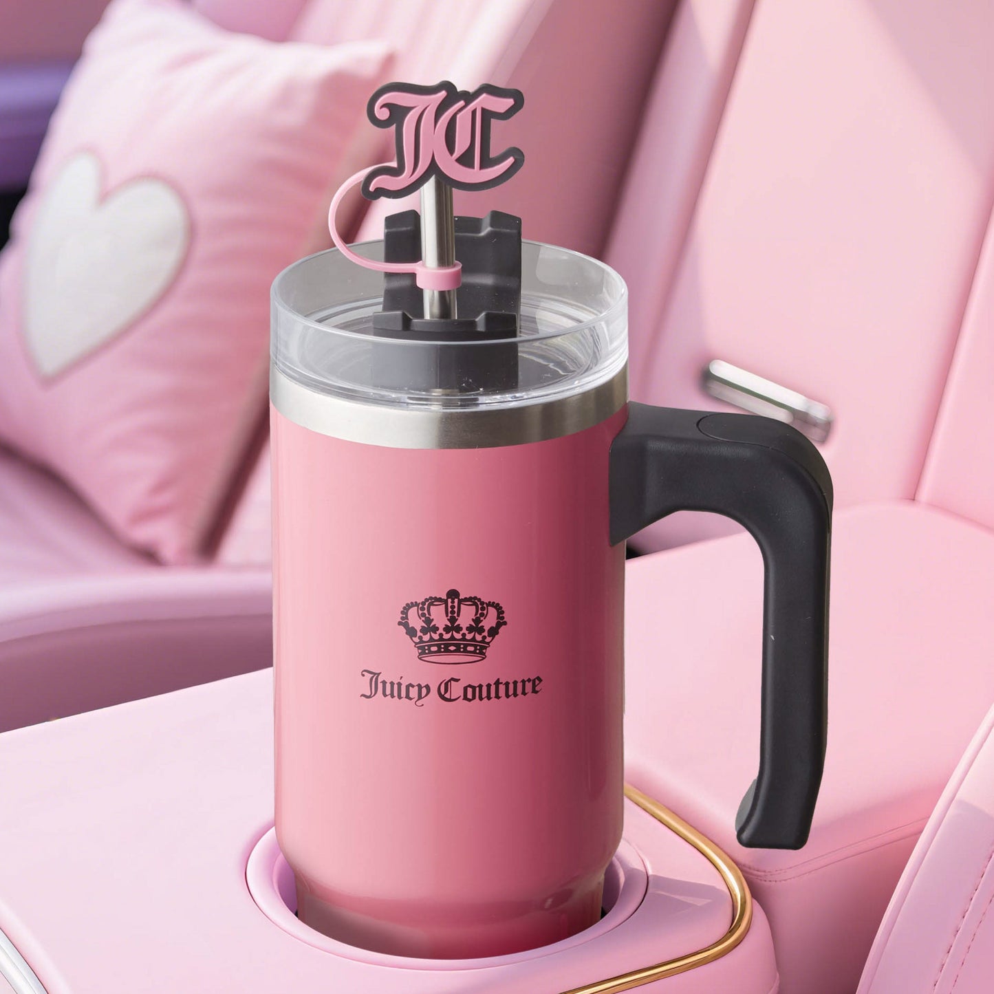 A large pink Jetsetter Hydration Stainless Steel Tumbler with a black handle sits in a car cup holder. It features a crown logo, “Juicy Couture” text, pink straw with “JC” topper, heart accents, and includes a neoprene crossbody pouch.