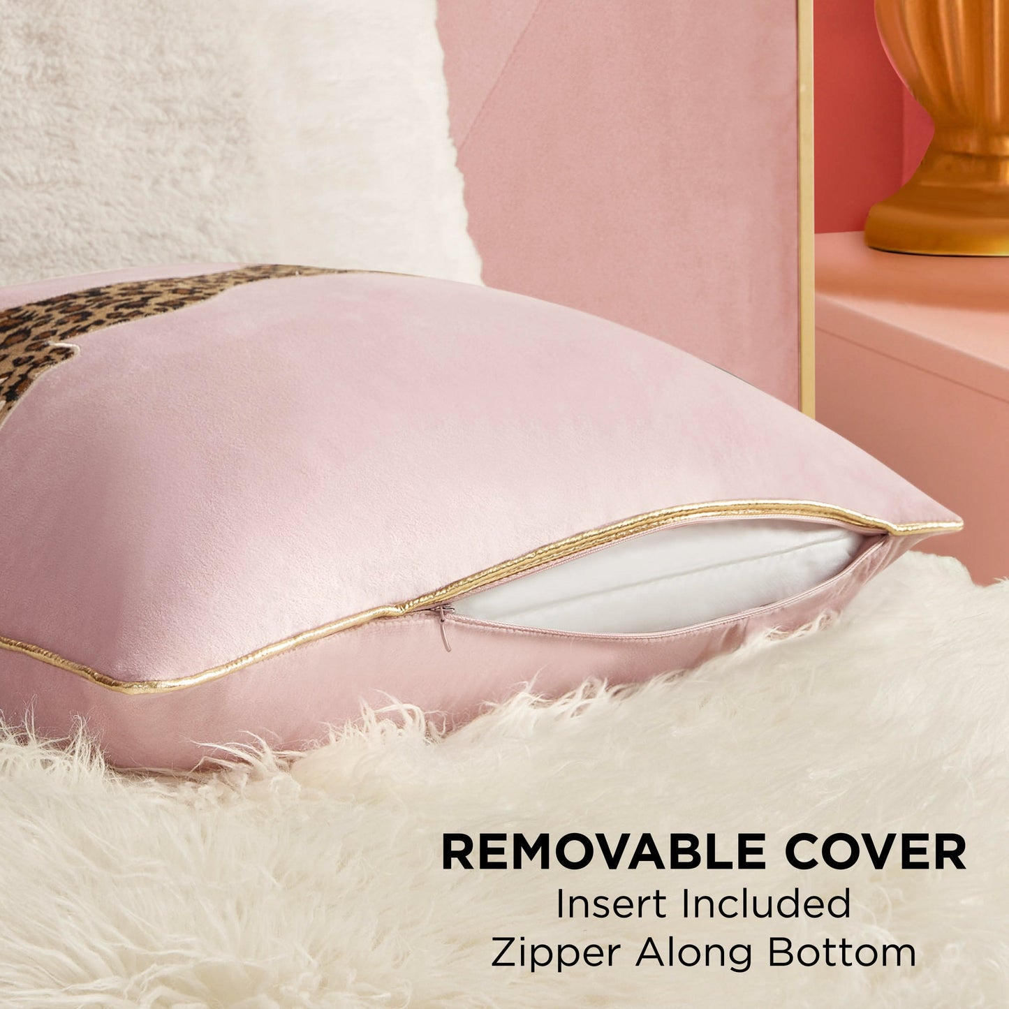 A close-up of the Velvet Cheetah Pillow shows its gold bottom zipper partially open to reveal the white insert. The removable cover rests on a fluffy surface, with a gold lamp and pink wall behind. Insert is included.