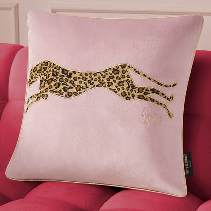 A square light pink Velvet Cheetah Pillow featuring an embroidered leaping cheetah with a leopard print sits on a red couch. It has gold piping, a side brand tag, and is offered by Juicy Couture as a decorative accent.