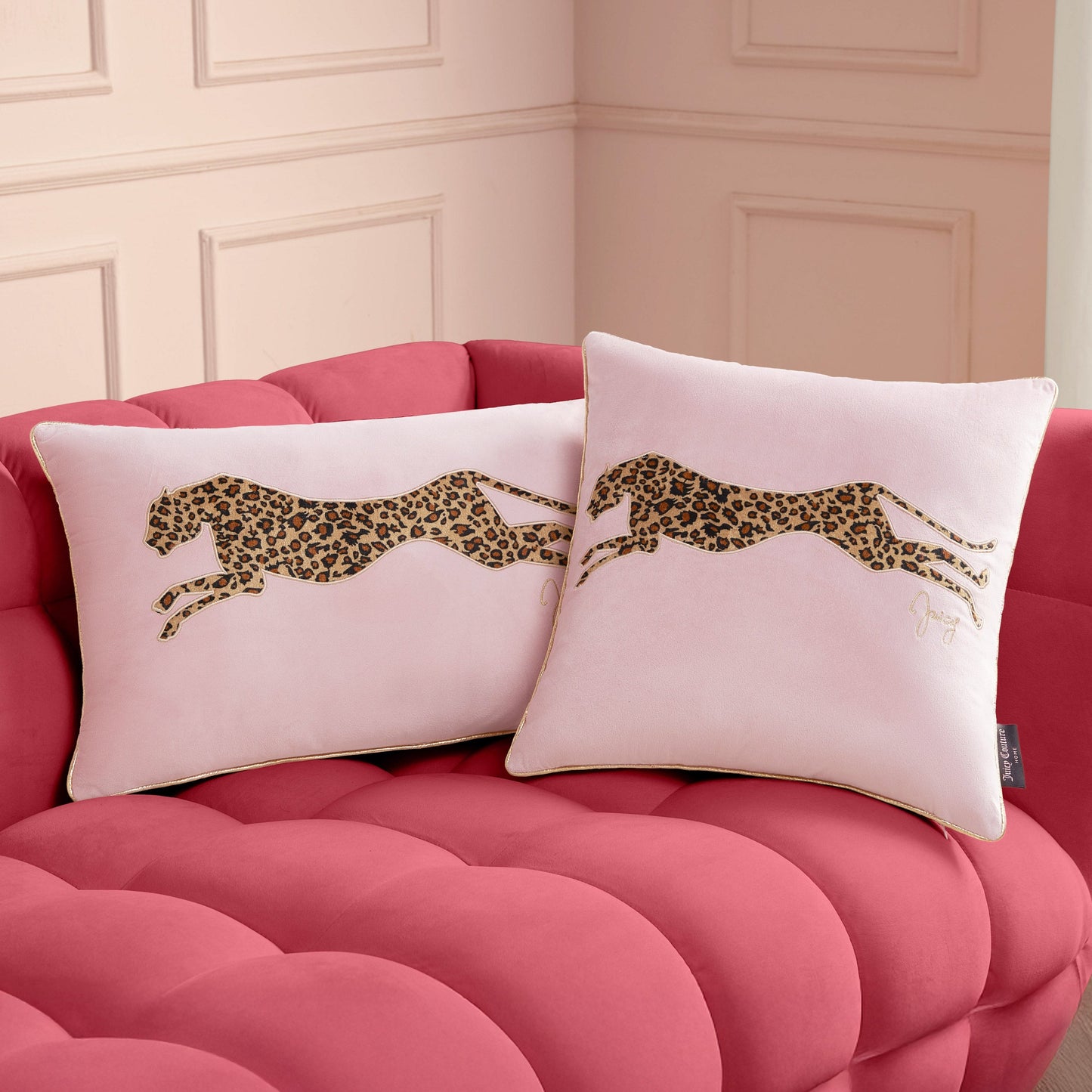 Two square pale pink Velvet Cheetah Pillows with embroidered cheetahs in a leopard print pattern rest on a tufted, bright pink velvet sofa. A light-colored wall with decorative molding is in the background.