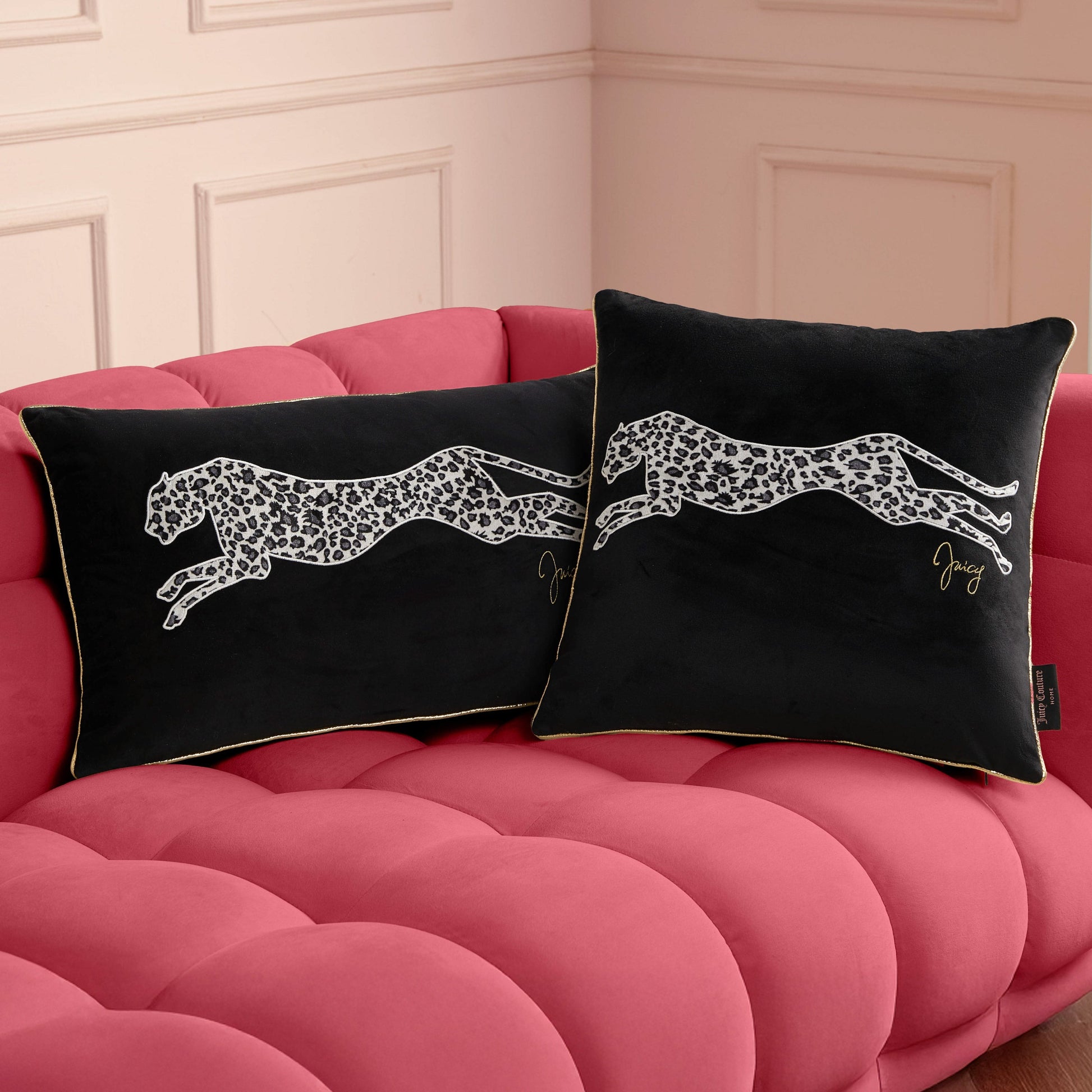 Two black Velvet Cheetah Square Pillows with white leaping cheetah designs and gold piping rest on a plush pink tufted sofa against a pale paneled wall.
