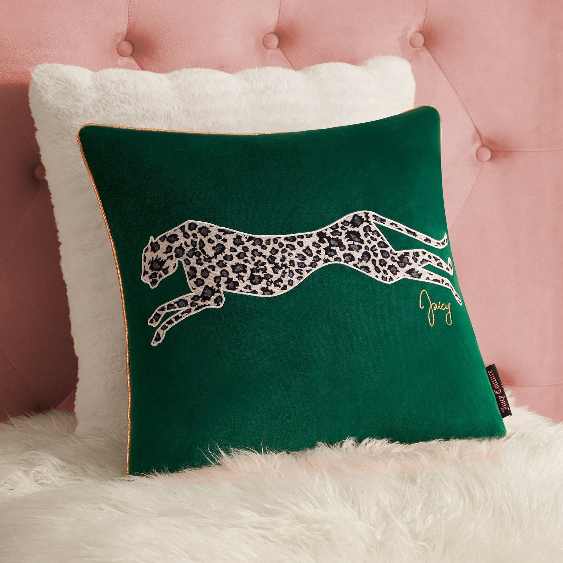 The Velvet Cheetah Square Pillow features a green velvet cover with a cheetah applique and “Juicy” in gold script, placed before a white fluffy pillow on a pink tufted background over white faux fur.