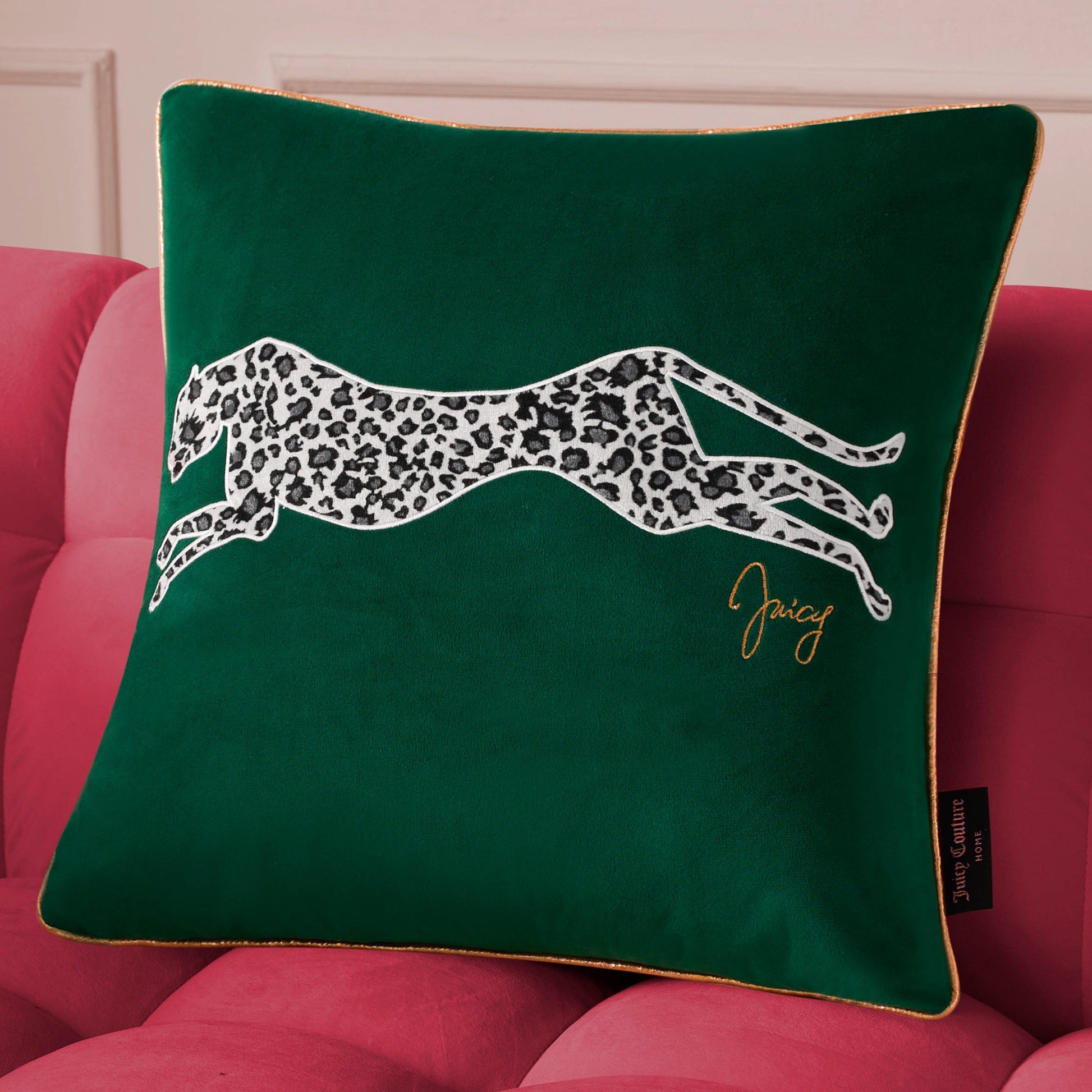 The Velvet Cheetah Square Pillow features a green velvet cover with a black-and-white cheetah applique and gold “Juicy” embroidery, styled on a pink couch for a chic Juicy Couture look.