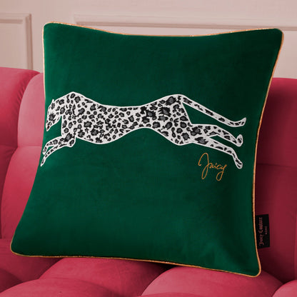 The Velvet Cheetah Square Pillow features a green velvet cover with a black-and-white cheetah applique and gold “Juicy” embroidery, styled on a pink couch for a chic Juicy Couture look.