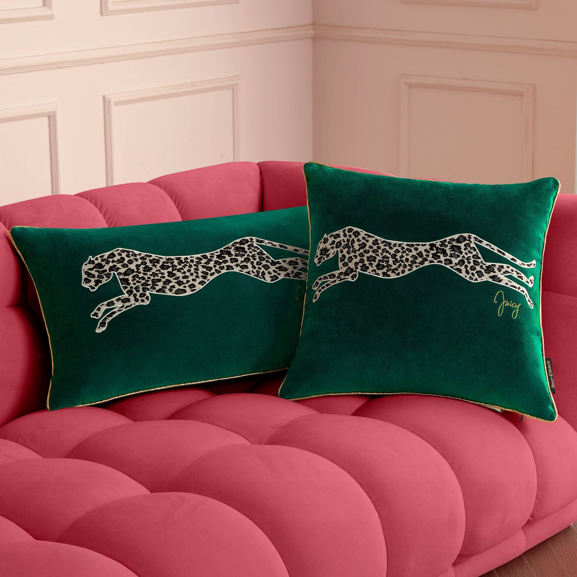 Two green Velvet Cheetah Square Pillows with bold cheetah appliques sit on a plush tufted pink sofa in front of a cream-colored wall with decorative molding.