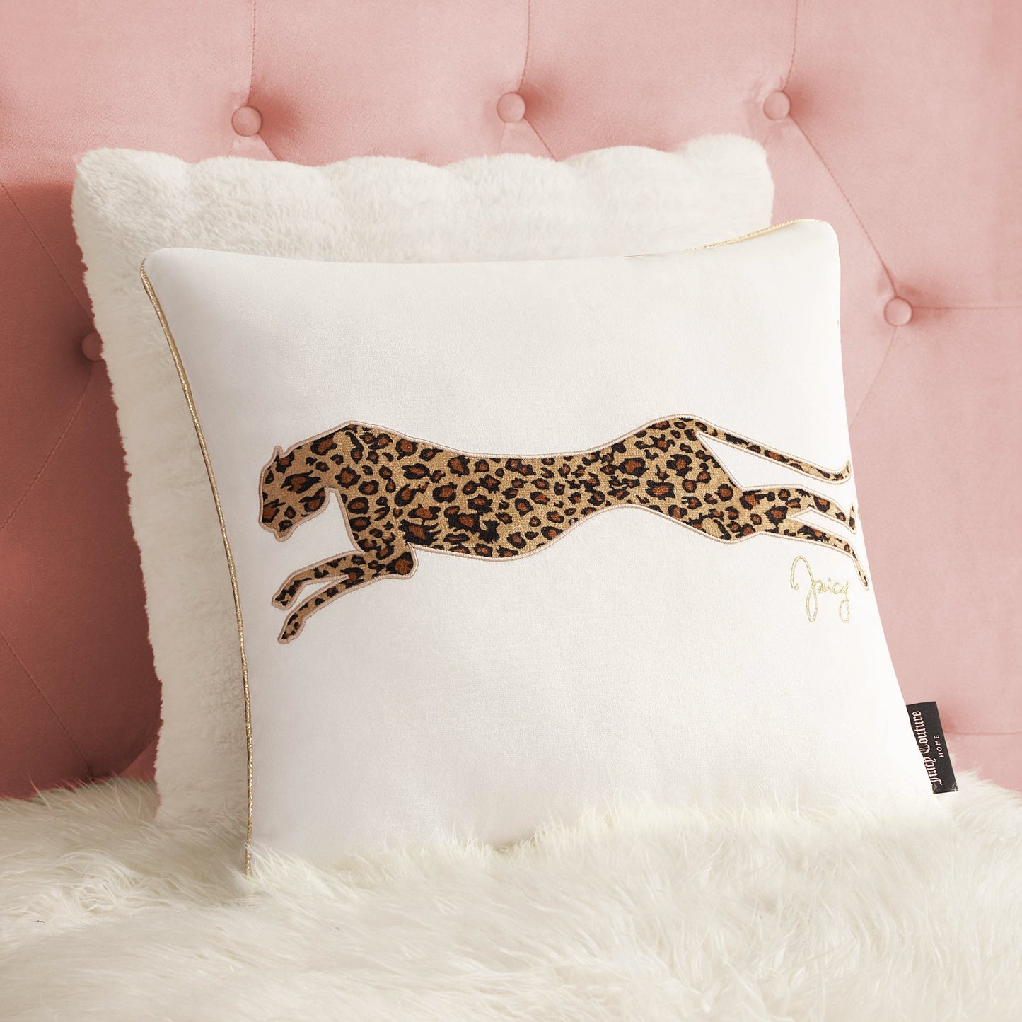 The Velvet Cheetah Pillow features a striking leaping leopard design with brown and black print, shown on a white furry surface against a pink tufted headboard—a bold accent for any room.