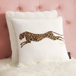 The Velvet Cheetah Pillow features a striking leaping leopard design with brown and black print, shown on a white furry surface against a pink tufted headboard—a bold accent for any room.