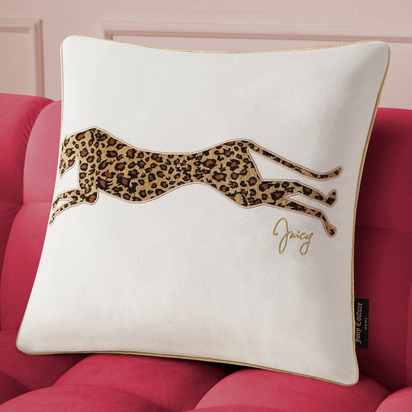 The Velvet Cheetah Pillow, featuring gold piping, an embroidered cheetah, and "Juicy" in gold script, adds a stylish Juicy Couture accent to any space when placed on a red tufted sofa.