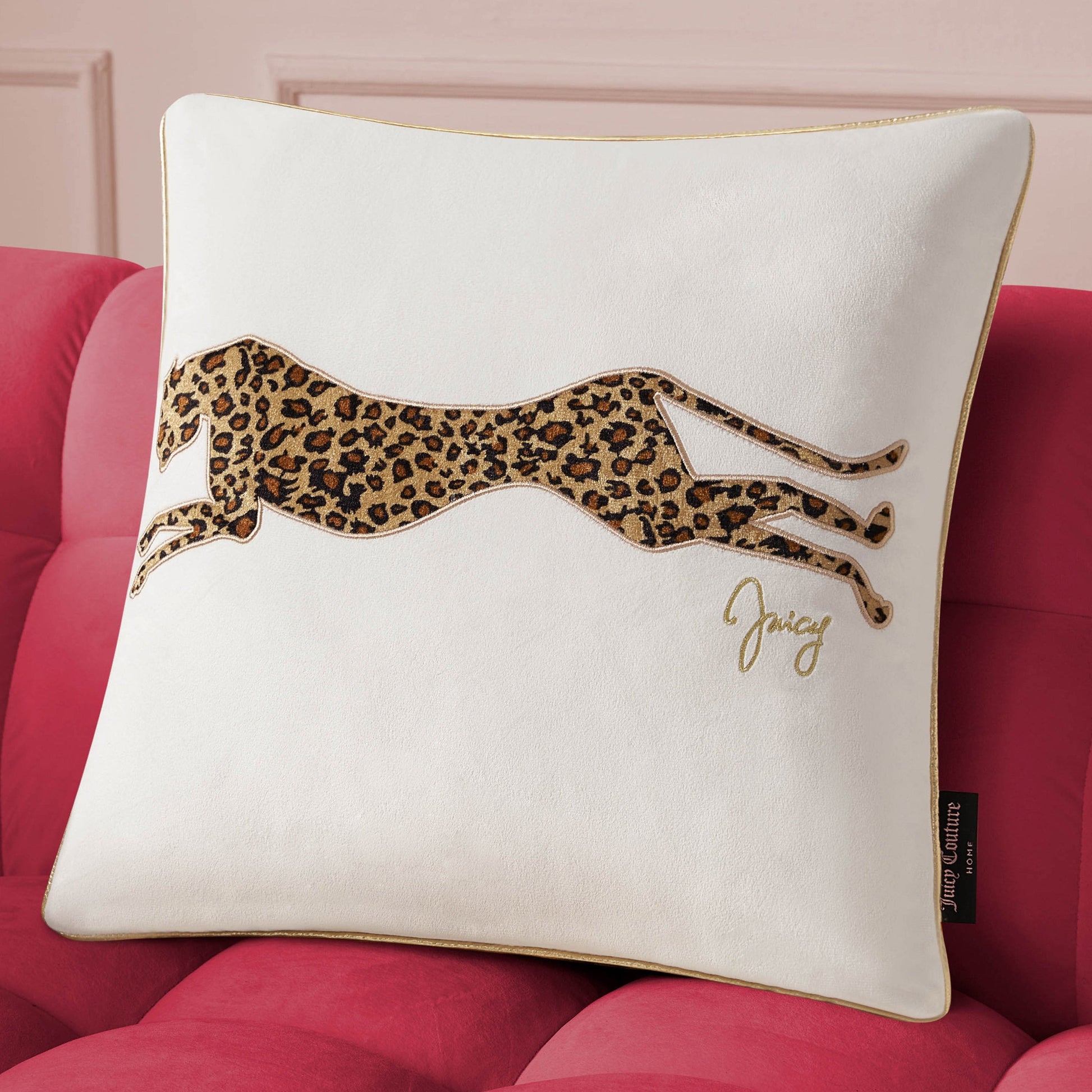 The Velvet Cheetah Pillow, featuring gold piping, an embroidered cheetah, and "Juicy" in gold script, adds a stylish Juicy Couture accent to any space when placed on a red tufted sofa.