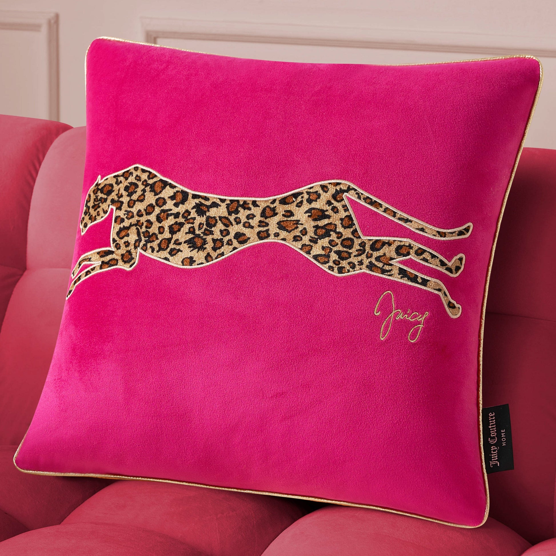 A bright pink Velvet Cheetah Square Pillow with a cheetah applique and "Juicy" in gold embroidery rests on a pink upholstered couch.