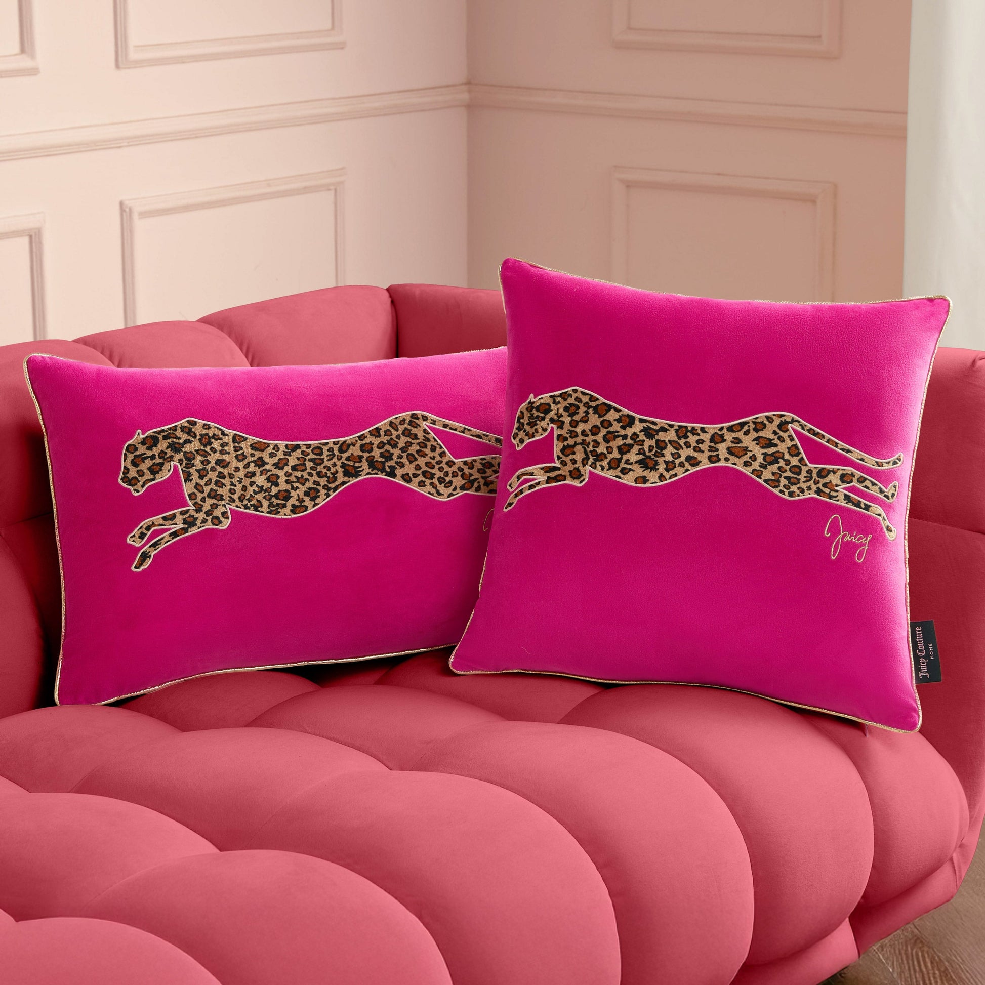 Two bright pink Velvet Cheetah Square Pillows with bold cheetah appliques rest on a plush, tufted pink sofa set against a light-colored paneled wall.