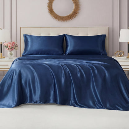 A neatly made bed features the Solid Satin Sheet Set in shiny navy blue with matching pillowcases, set against a light tufted headboard. Bedside tables hold decorative lamps and flowers, while a round mirror hangs above the bed.