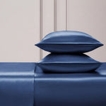 Two shiny blue satin pillows are stacked on the Solid Satin Sheet Set, all in a deep blue shade, set against a light textured wall with simple paneling—a scene that evokes Juicy Couture's signature luxe style.