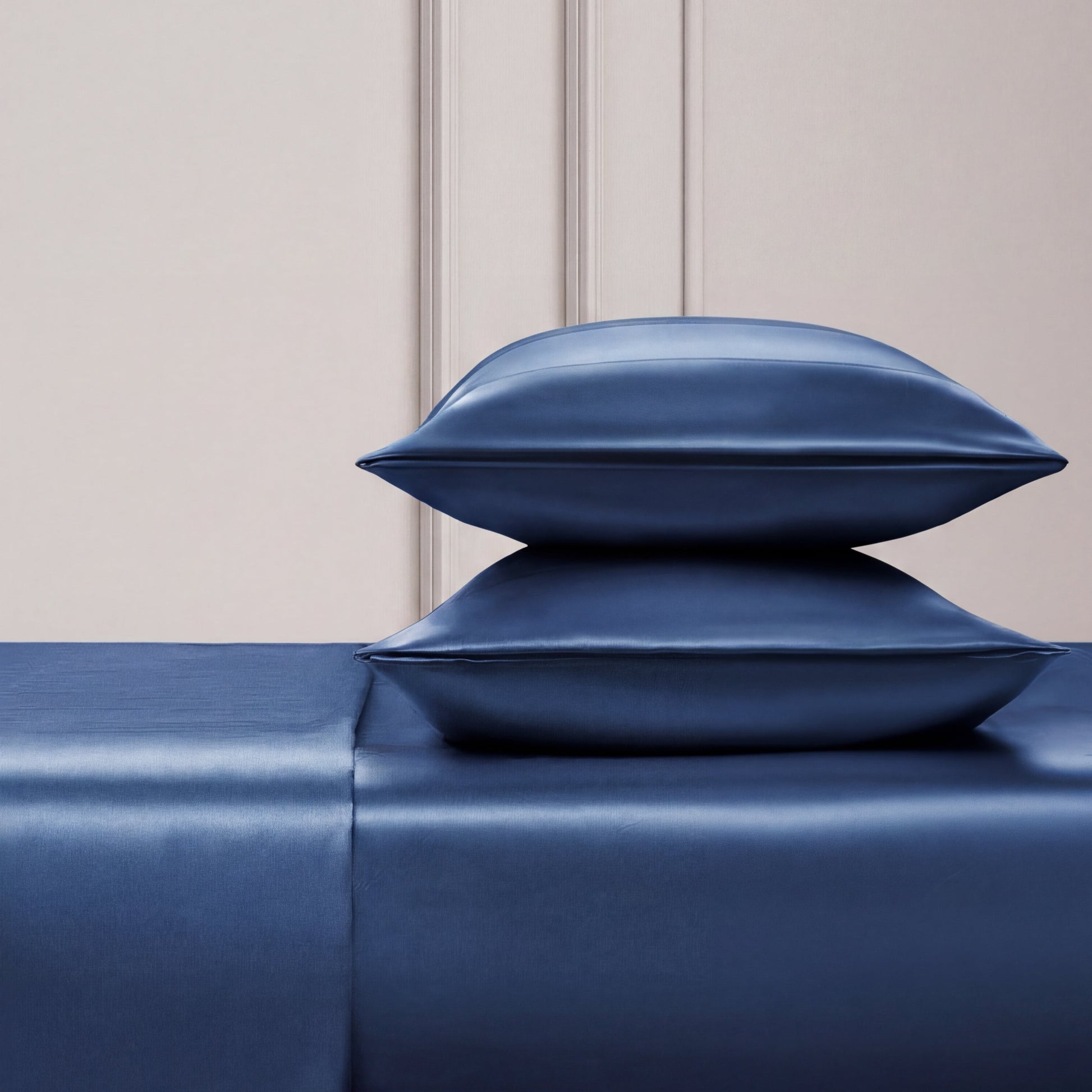 Two shiny blue satin pillows are stacked on the Solid Satin Sheet Set, all in a deep blue shade, set against a light textured wall with simple paneling—a scene that evokes Juicy Couture's signature luxe style.