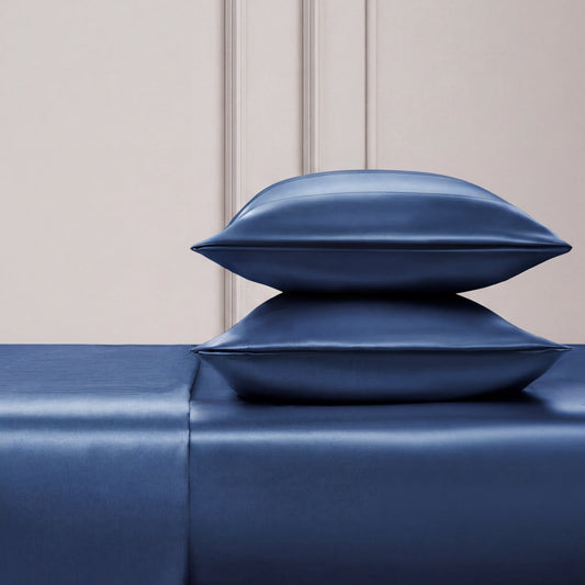Two shiny blue satin pillows are stacked on the Solid Satin Sheet Set, all in a deep blue shade, set against a light textured wall with simple paneling—a scene that evokes Juicy Couture's signature luxe style.