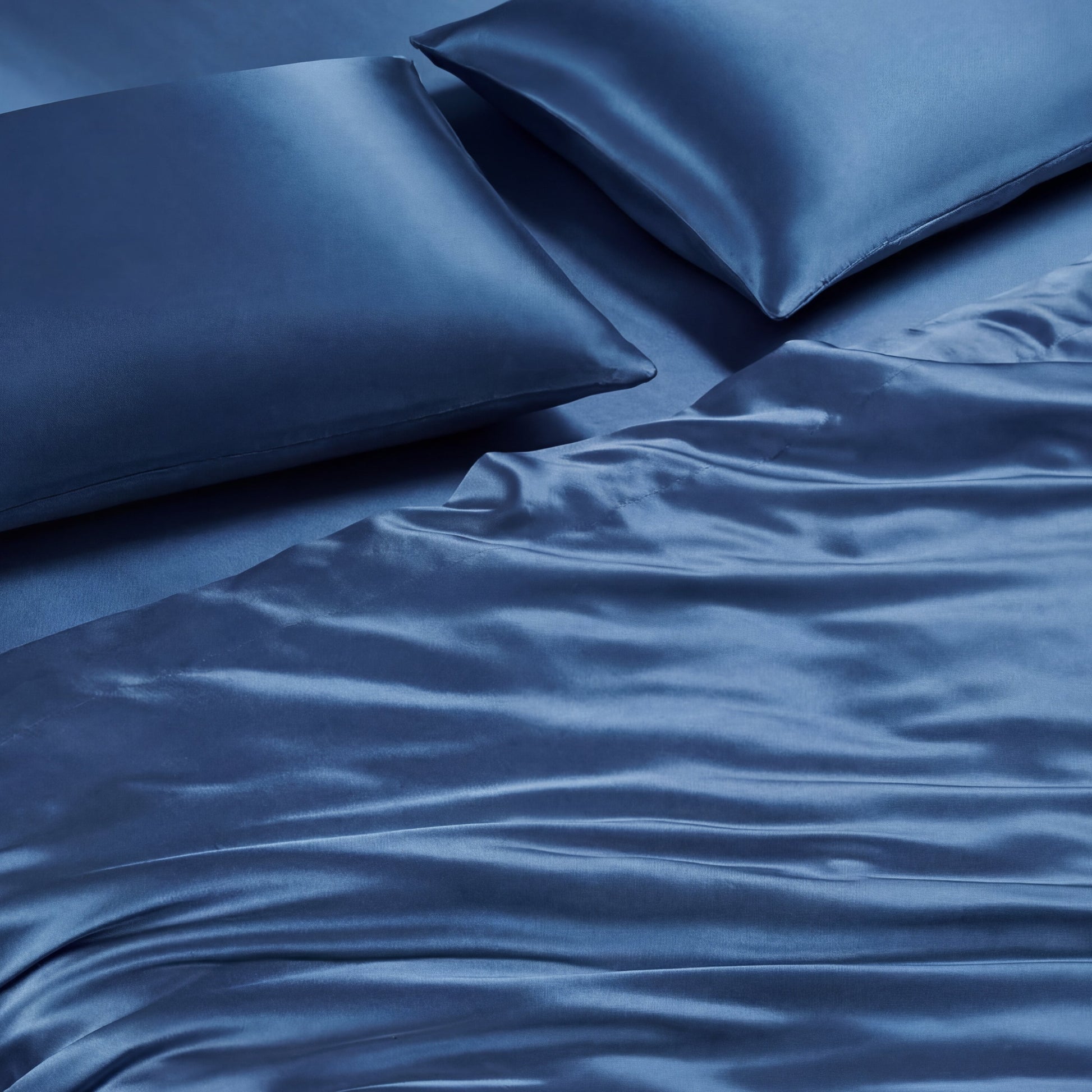 Two blue Solid Satin Sheet Set pillows and a matching ultra-soft satin sheet set with a smooth, shiny texture are arranged on a bed, creating a luxurious and calming appearance.