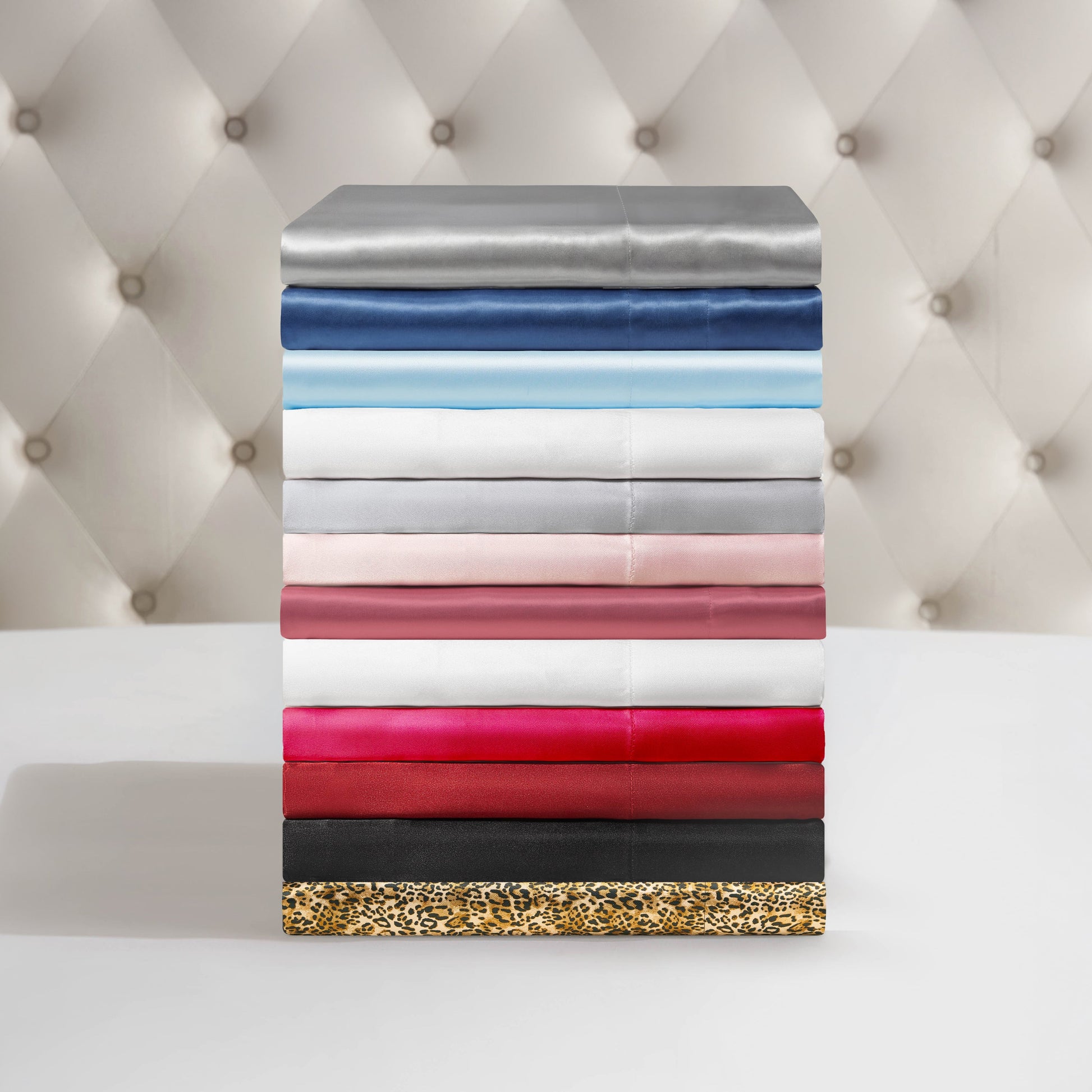 A neatly stacked pile of Solid Satin Sheet Sets in assorted colors and patterns—grey, blue, white, pink, red, black, and leopard print—displayed against a tufted beige background.