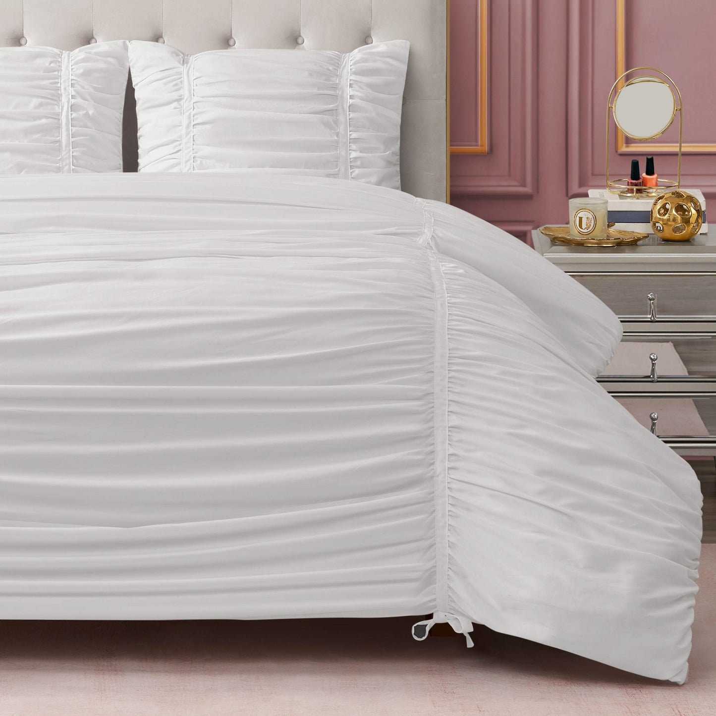 A neatly made bed with the Ribbon Ruched Comforter Set and matching pillows stands by a silver nightstand holding a mirror, perfume bottles, and gold décor, set against a mauve paneled wall.