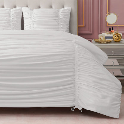 A neatly made bed with the Ribbon Ruched Comforter Set and matching pillows stands by a silver nightstand holding a mirror, perfume bottles, and gold décor, set against a mauve paneled wall.
