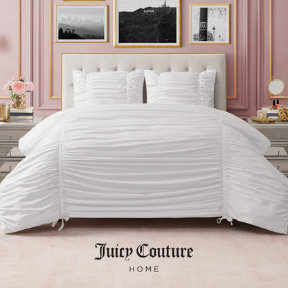 A stylish bedroom features a neatly made bed with the Ribbon Ruched Comforter Set and pillows, pink walls, gold-framed black-and-white photos, elegant bedside tables, and "Juicy Couture Home" at the bottom.