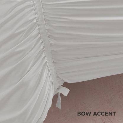 Close-up of the Ribbon Ruched Comforter Set in white, featuring ribbon detailing and a side bow accent, draped over an edge with a pink floor beneath. "BOW ACCENT" appears in black text at the lower right corner.