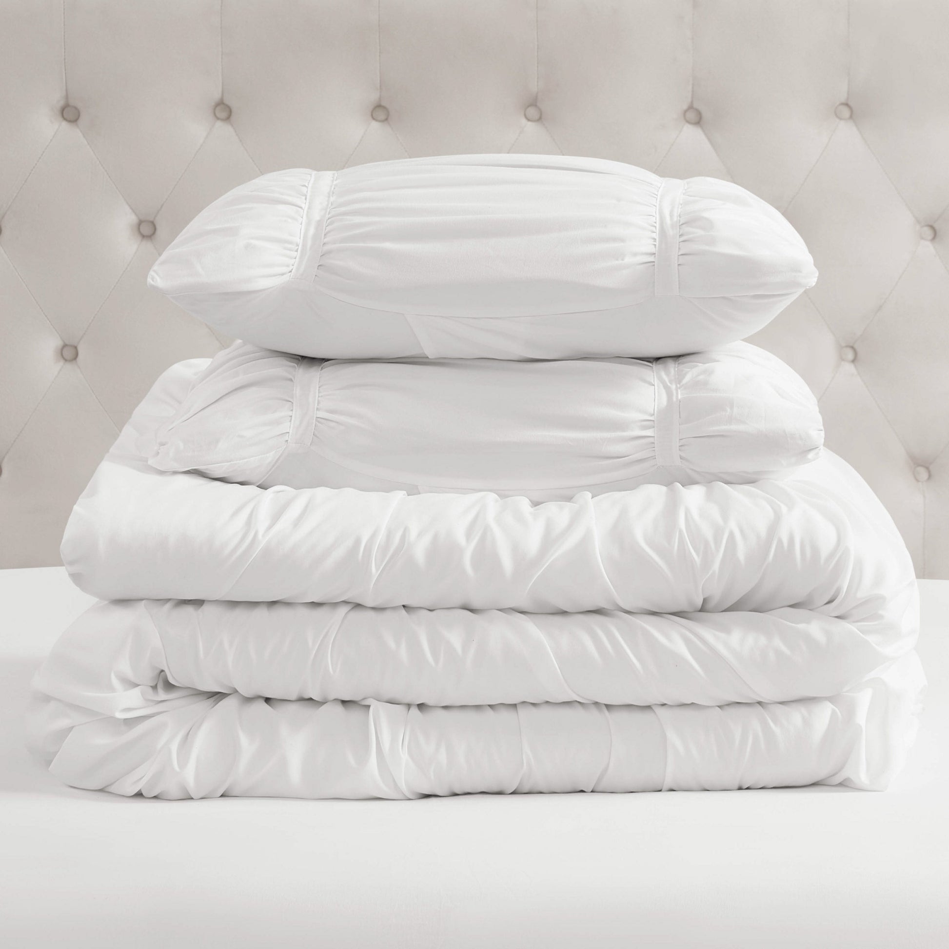 A stack of white bedding, featuring two pillows and the Ribbon Ruched Comforter Set, is neatly arranged on a bed with a tufted, light-colored headboard in the background.