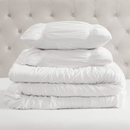 A stack of white bedding, featuring two pillows and the Ribbon Ruched Comforter Set, is neatly arranged on a bed with a tufted, light-colored headboard in the background.