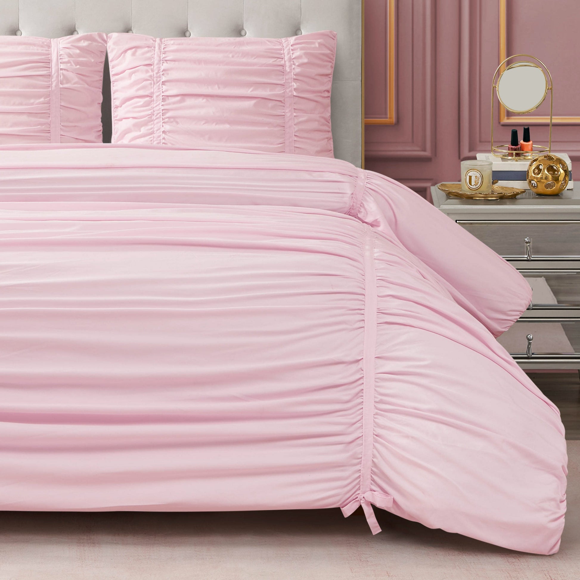 A neatly made bed with a light pink Ribbon Ruched Comforter Set and matching pillows sits beside a silver nightstand topped with a round mirror, perfume bottles, and decor. Elegant wall molding enhances the room’s stylish look.