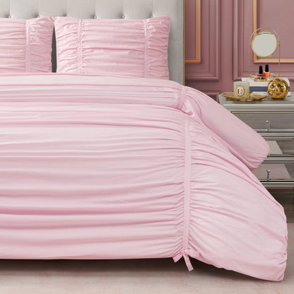 A neatly made bed with a light pink Ribbon Ruched Comforter Set and matching pillows sits beside a silver nightstand topped with a round mirror, perfume bottles, and decor. Elegant wall molding enhances the room’s stylish look.