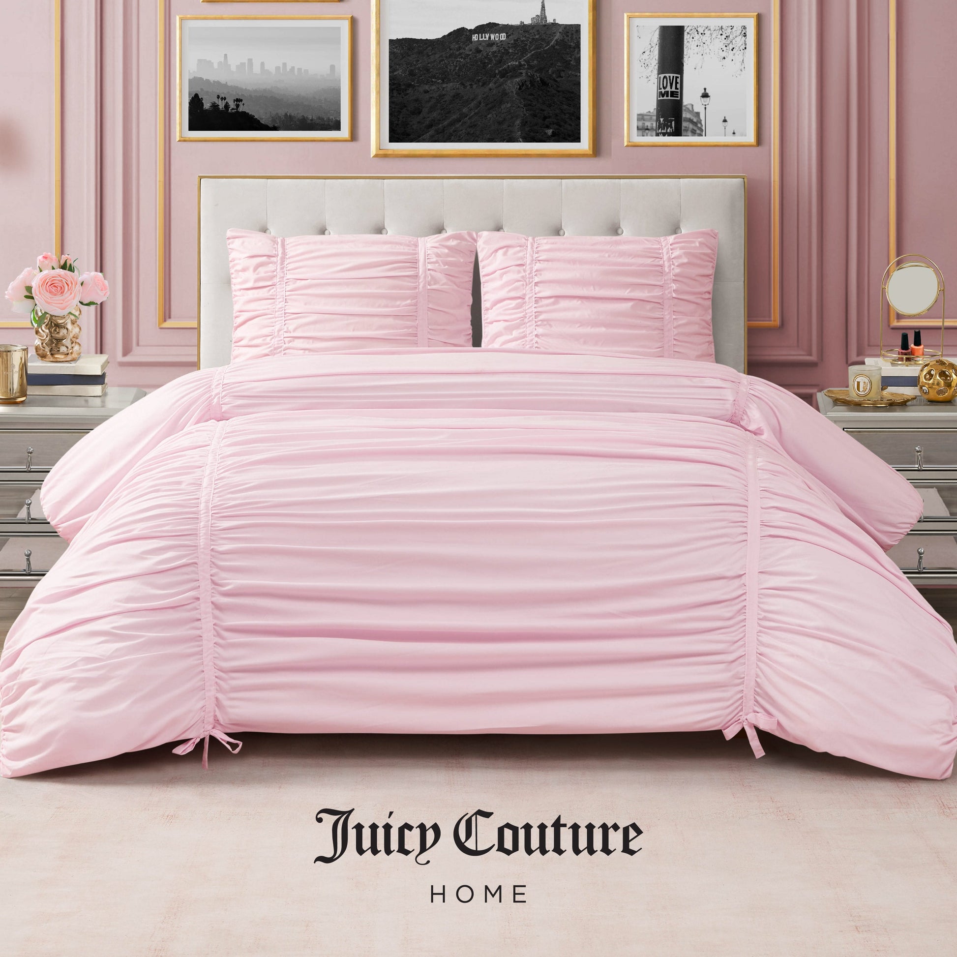 A bedroom features a tufted beige headboard, a neatly made bed with the Ribbon Ruched Comforter Set by Juicy Couture Home in pale pink, matching pillows, silver nightstands, flowers, and framed art for an elegant touch. "Juicy Couture Home" logo at the bottom.