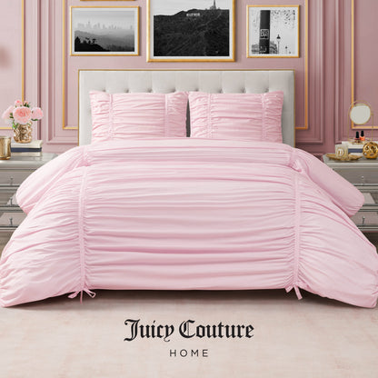 A bedroom features a tufted beige headboard, a neatly made bed with the Ribbon Ruched Comforter Set by Juicy Couture Home in pale pink, matching pillows, silver nightstands, flowers, and framed art for an elegant touch. "Juicy Couture Home" logo at the bottom.