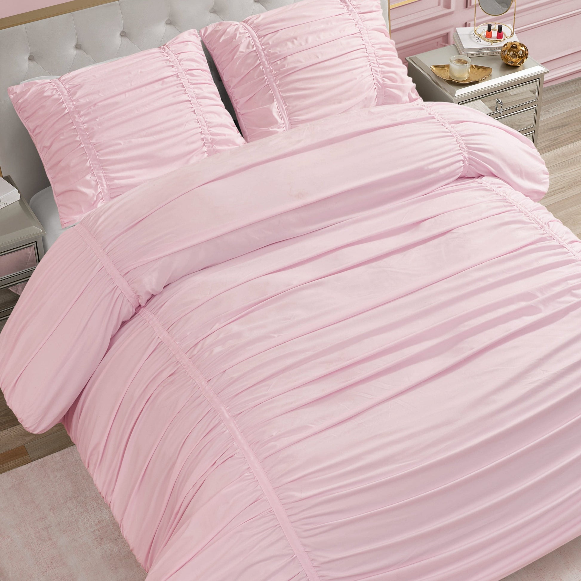 A neatly made bed featuring the Ribbon Ruched Comforter Set in light pink with matching pillows, placed in a bedroom with light furniture and a silver nightstand displaying a tray with a teapot, cup, and pastries.