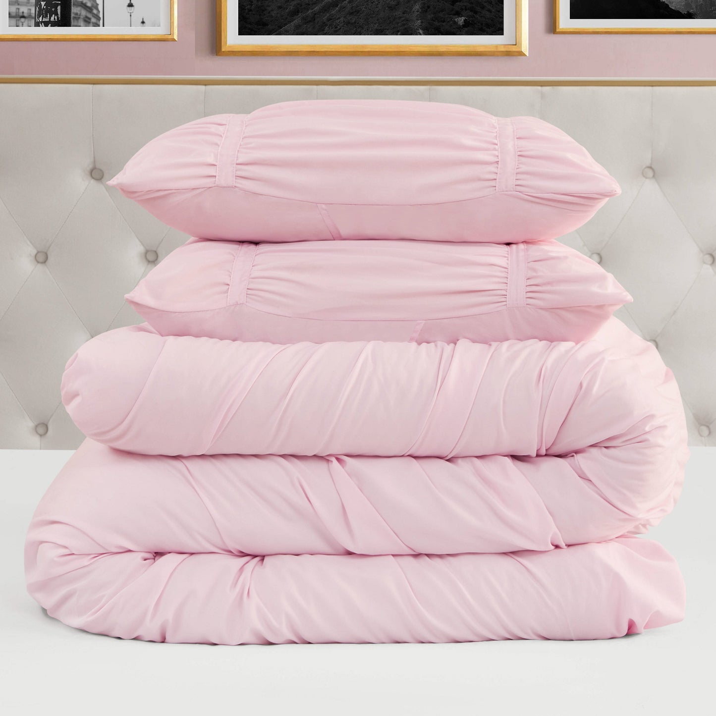 A neatly stacked Ribbon Ruched Comforter Set in light pink, featuring two pillows and two folded comforters with ribbon detailing, is placed on a white surface in front of a tufted headboard and framed wall art.