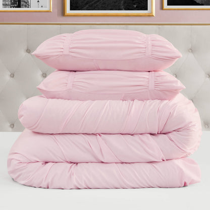 A neatly stacked Ribbon Ruched Comforter Set in light pink, featuring two pillows and two folded comforters with ribbon detailing, is placed on a white surface in front of a tufted headboard and framed wall art.