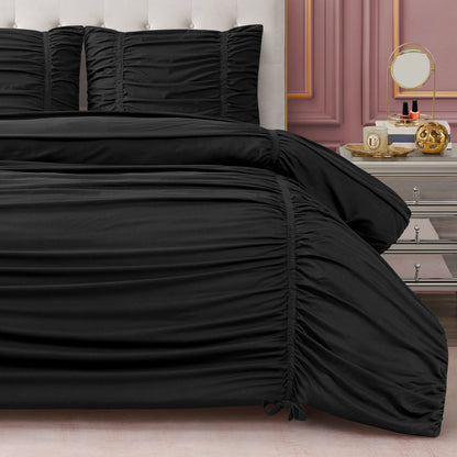 A bed with a black Ribbon Ruched Comforter Set featuring a ruched comforter and shams sits against a tufted headboard. A mirrored nightstand displays perfume in a pink-walled room accented with chic ribbon details.