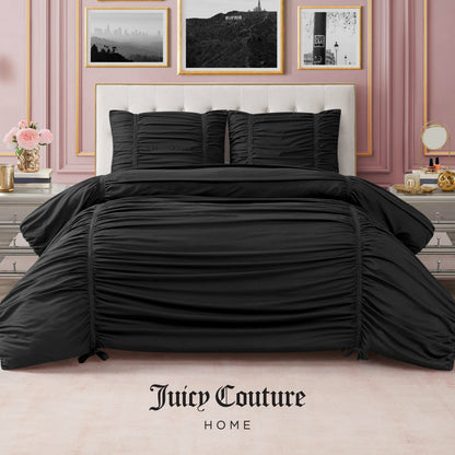 A stylish bedroom with pink walls has a neatly made bed featuring the Ribbon Ruched Comforter Set in black, matching pillows, gold-framed art, and decorative items on mirrored nightstands. Juicy Couture Home is printed on the floor.